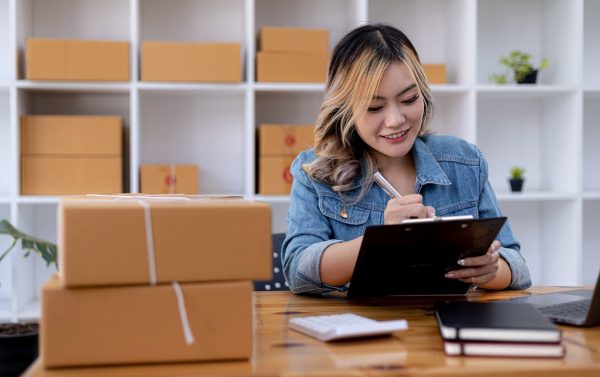 A Young Lady Appears To Be In An Office With Packages  Behind Her And On The Table In Front Of Her And She Appears To Also Have A Pen In Her Hands While  Holding What Looks Like A Check List.