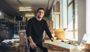 Successful Online Business Owner Sitting At His Workdesk. Young Man With Laptop And Shipment Box On Desk, Looking At Camera And Smiling.