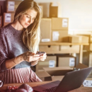 A young lady standing at her desk with packages around her while looking at her phone to calculate her shipping cost.
